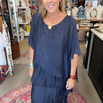 A woman wearing a navy blue tiered tunic maxi dress stands inside a boutique shop. The dress has a loose, draped fit with short sleeves and a V-neckline.