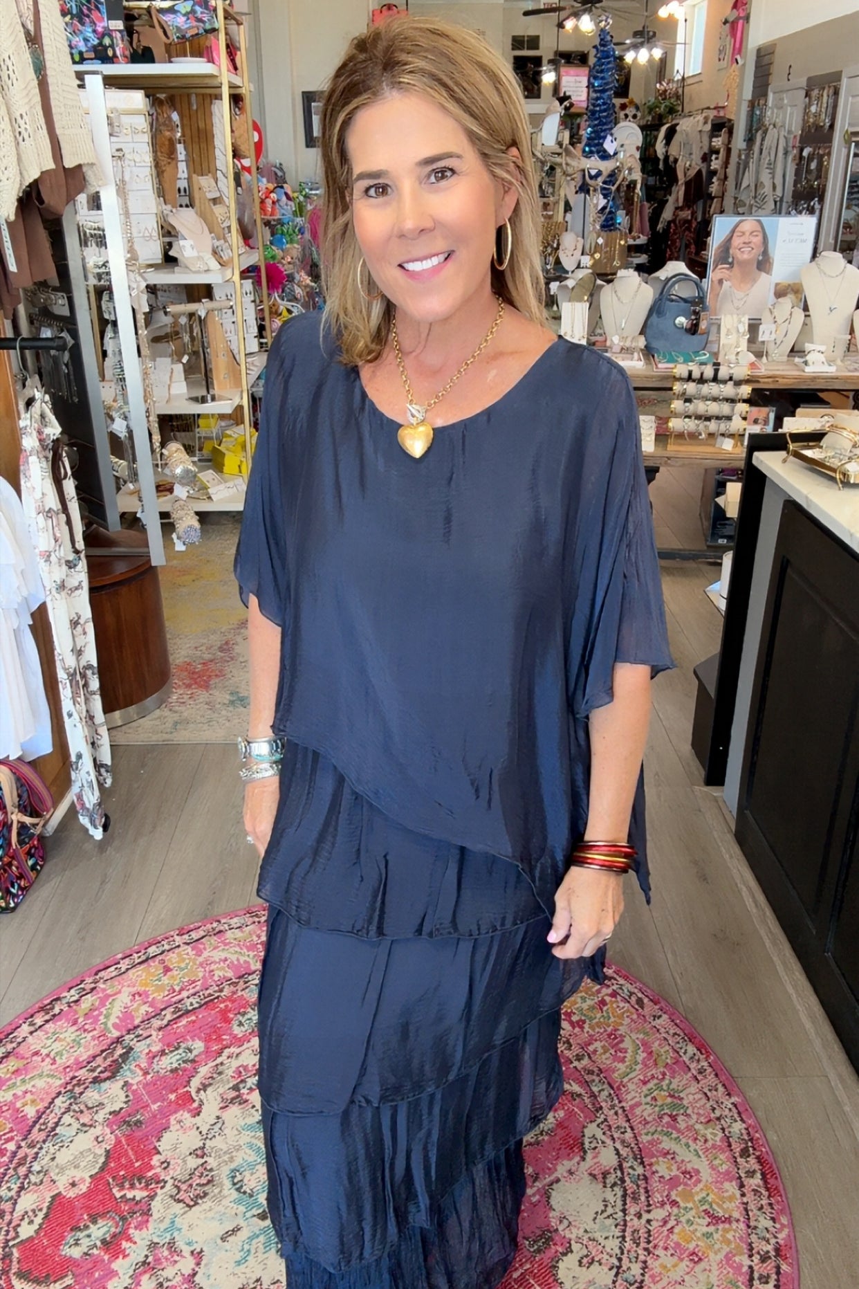 A woman wearing a navy blue tiered tunic maxi dress stands inside a boutique shop. The dress has a loose, draped fit with short sleeves and a V-neckline.