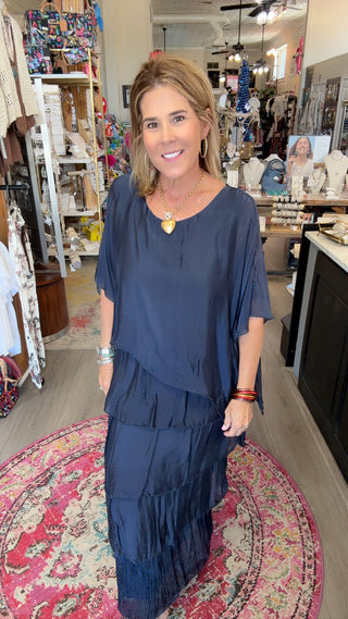 A woman wearing a navy blue tiered tunic maxi dress stands inside a boutique shop. The dress has a loose, draped fit with short sleeves and a V-neckline.