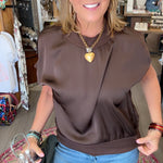 A woman wearing a brown short-sleeve top and blue jeans stands inside a clothing boutique, holding a black garment bag. She is wearing a gold heart pendant necklace.