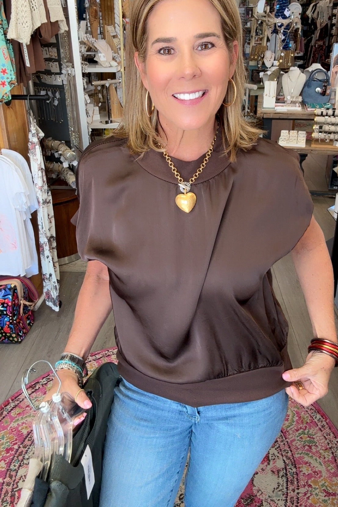 A woman wearing a brown short-sleeve top and blue jeans stands inside a clothing boutique, holding a black garment bag. She is wearing a gold heart pendant necklace.