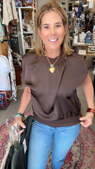 A woman wearing a brown short-sleeve top and blue jeans stands inside a clothing boutique, holding a black garment bag. She is wearing a gold heart pendant necklace.