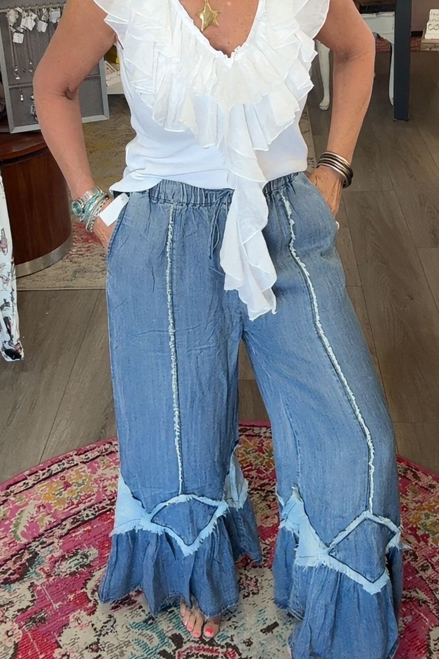 A woman stands barefoot on a patterned rug inside a boutique, wearing a white ruffled sleeveless top and wide-leg blue jeans with light blue trim.