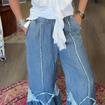 A woman stands barefoot on a patterned rug inside a boutique, wearing a white ruffled sleeveless top and wide-leg blue jeans with light blue trim.