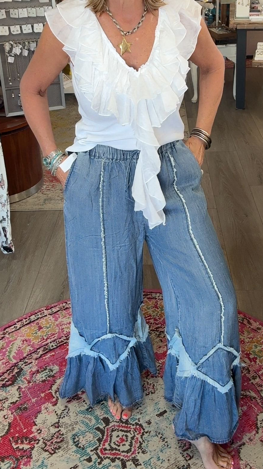 A woman stands barefoot on a patterned rug inside a boutique, wearing a white ruffled sleeveless top and wide-leg blue jeans with light blue trim.