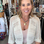 A woman smiling in a boutique, wearing a cream open-knit cardigan over a light pink top and beige pants. The background shows a retail store interior with clothing racks and displays.