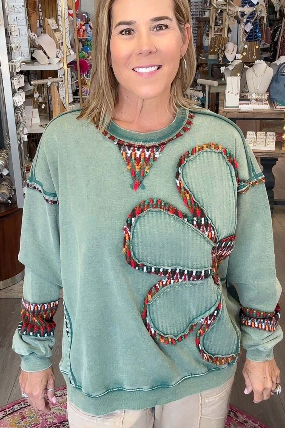 A woman modeling a sage green long-sleeve sweater with a large butterfly design outlined in colorful beads, paired with cream pants, standing inside a retail store.