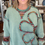 A woman modeling a sage green long-sleeve sweater with a large butterfly design outlined in colorful beads, paired with cream pants, standing inside a retail store.
