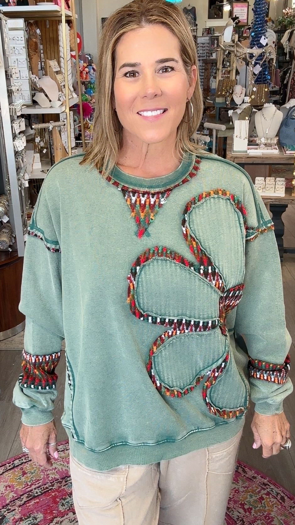 A woman modeling a sage green long-sleeve sweater with a large butterfly design outlined in colorful beads, paired with cream pants, standing inside a retail store.