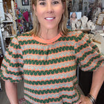 A woman wearing a short-sleeve knit top with green, orange, and white wavy stripes, paired with light beige pants, standing in a retail store.