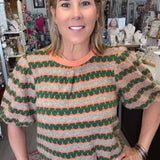 A woman wearing a short-sleeve knit top with green, orange, and white wavy stripes, paired with light beige pants, standing in a retail store.