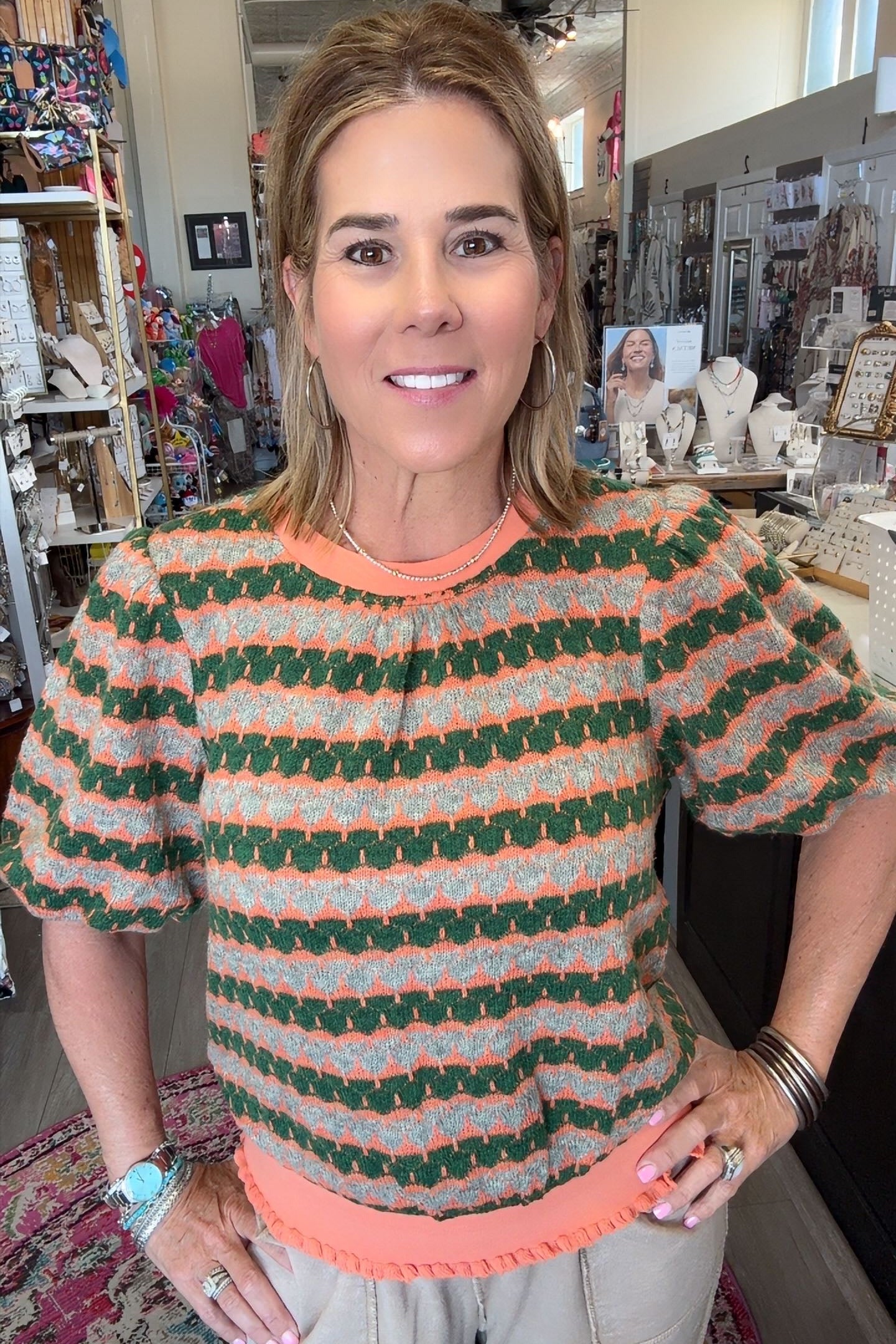 A woman wearing a short-sleeve knit top with green, orange, and white wavy stripes, paired with light beige pants, standing in a retail store.