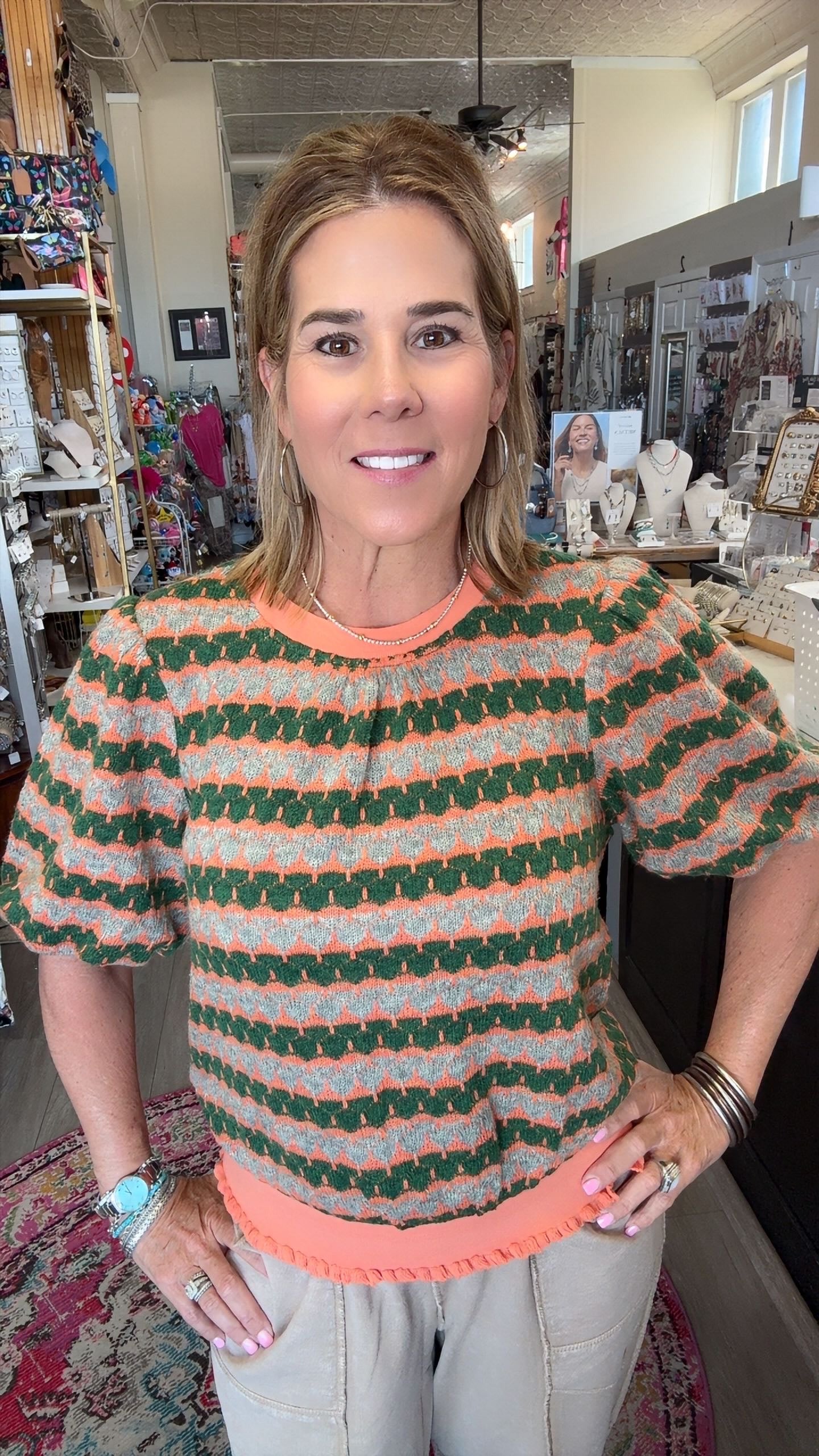 A woman wearing a short-sleeve knit top with green, orange, and white wavy stripes, paired with light beige pants, standing in a retail store.