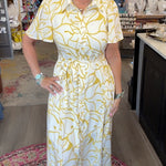 A woman modeling a white short-sleeve midi dress with a yellow abstract pattern, standing inside a retail boutique.