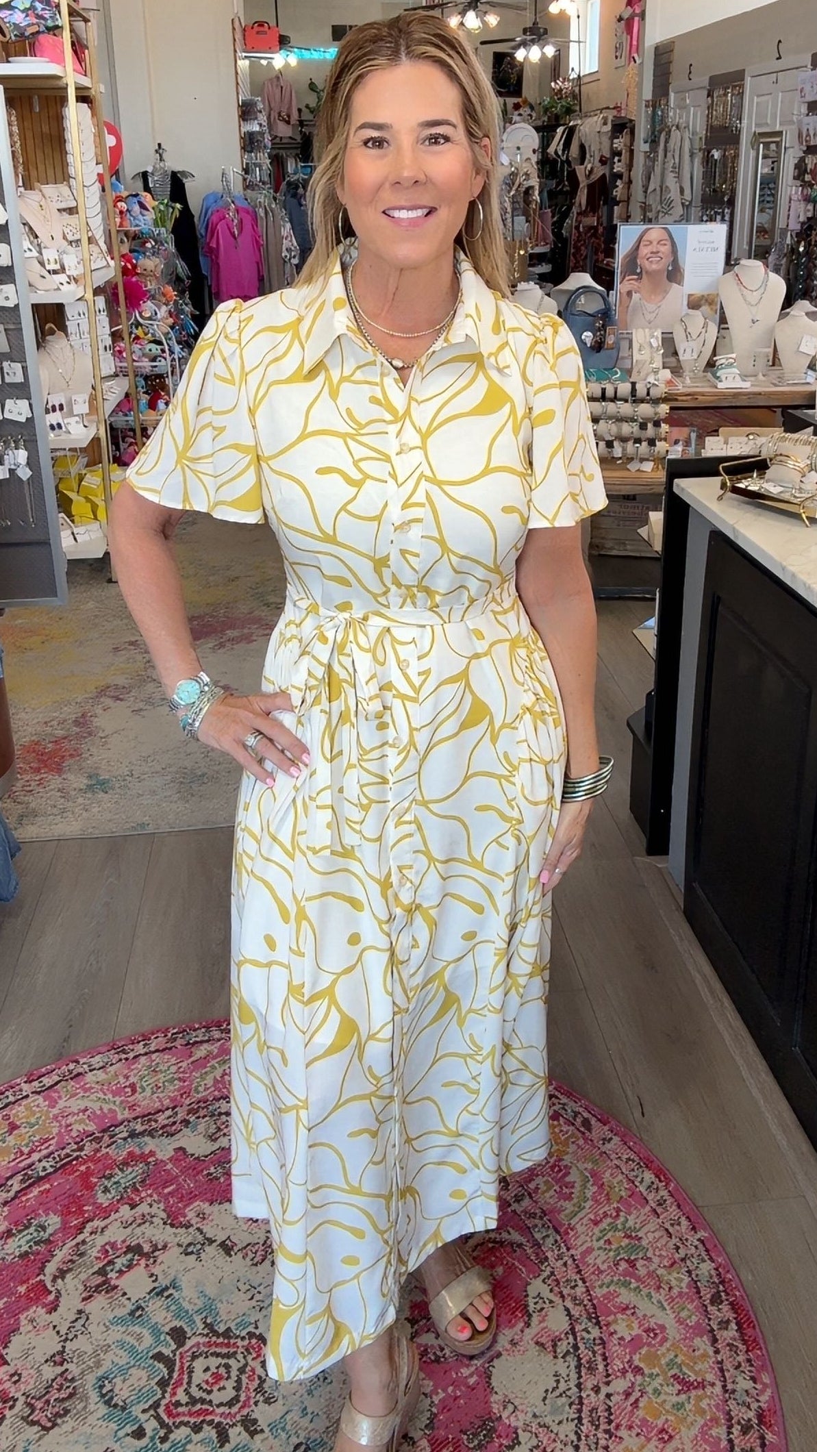 A woman modeling a white short-sleeve midi dress with a yellow abstract pattern, standing inside a retail boutique.