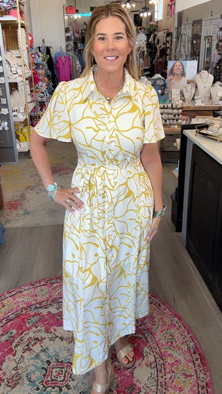 A woman modeling a white short-sleeve midi dress with a yellow abstract pattern, standing inside a retail boutique.