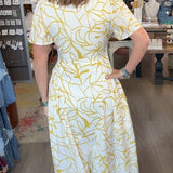 A woman wearing a white short-sleeve dress with a yellow abstract leaf pattern stands in a boutique, viewed from behind as she looks at her reflection in a mirror. The shop interior features a large pink patterned rug, wooden floors, and various clothing racks in the background.