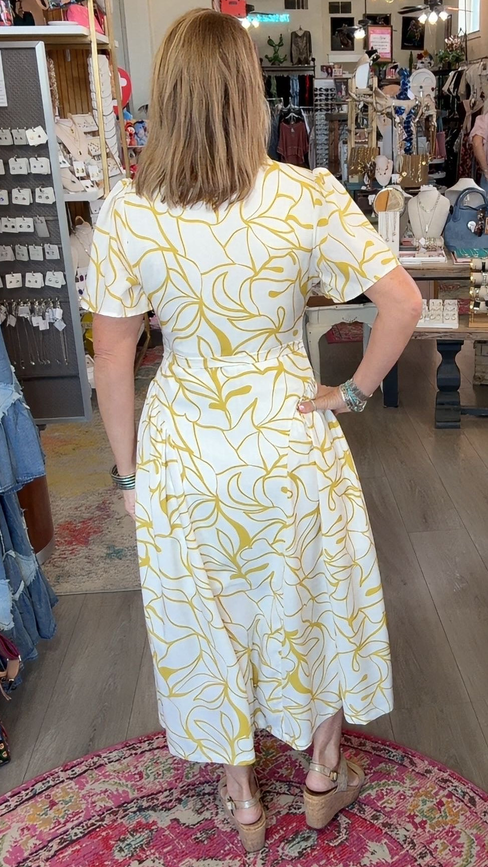 A woman wearing a white short-sleeve dress with a yellow abstract leaf pattern stands in a boutique, viewed from behind as she looks at her reflection in a mirror. The shop interior features a large pink patterned rug, wooden floors, and various clothing racks in the background.