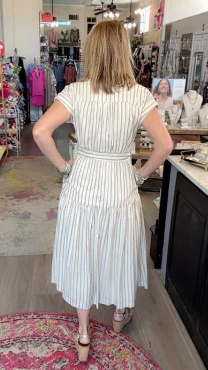 A woman wearing a white and light blue vertically striped midi dress, viewed from behind as she stands inside a boutique store.