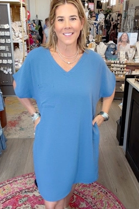 A woman wearing a light blue short-sleeve V-neck dress stands inside a retail boutique, smiling at the camera. The shop has a patterned rug, clothing racks, and display tables in the background.