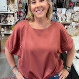 A woman with blonde hair stands in a boutique shop wearing a dusty rose short-sleeve top and blue jeans.