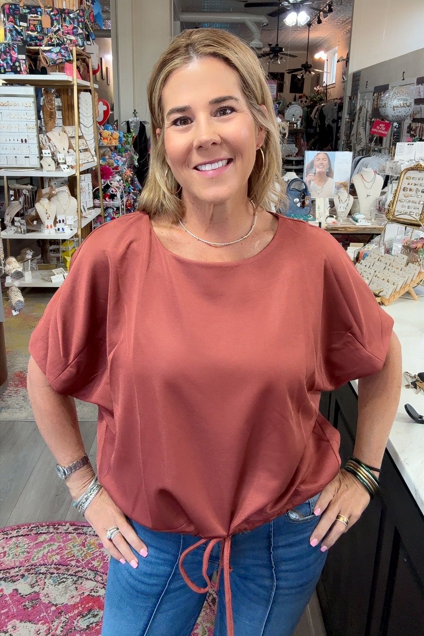 A woman with blonde hair stands in a boutique shop wearing a dusty rose short-sleeve top and blue jeans.