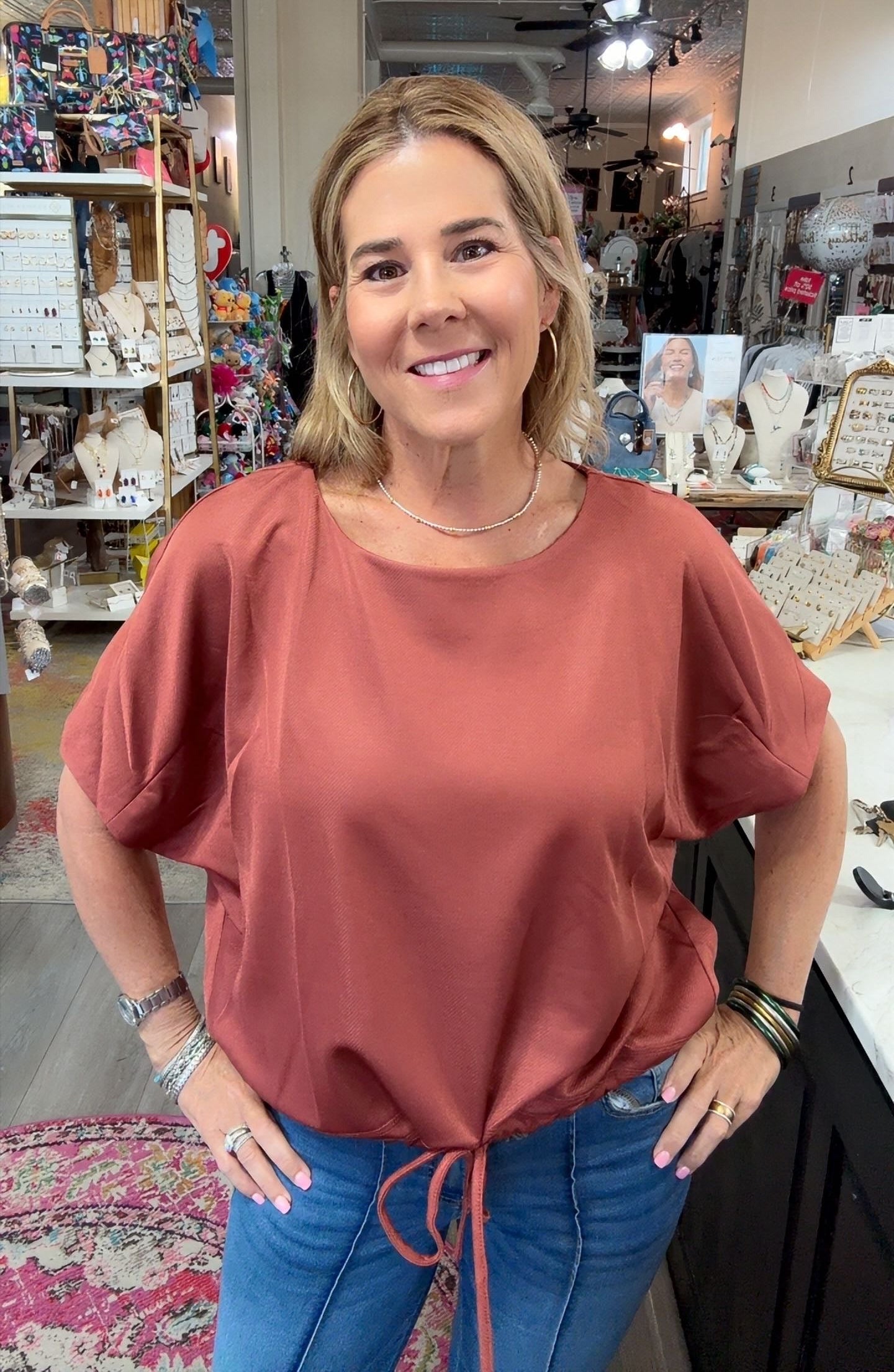 A woman with blonde hair stands in a boutique shop wearing a dusty rose short-sleeve top and blue jeans.