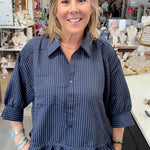 A woman with blonde hair smiling in a boutique shop, wearing a dark navy blue and black vertically striped button-down shirt with ruffle details at the sleeves and hem, paired with blue jeans.