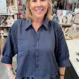 A woman with blonde hair smiling in a boutique shop, wearing a dark navy blue and black vertically striped button-down shirt with ruffle details at the sleeves and hem, paired with blue jeans.