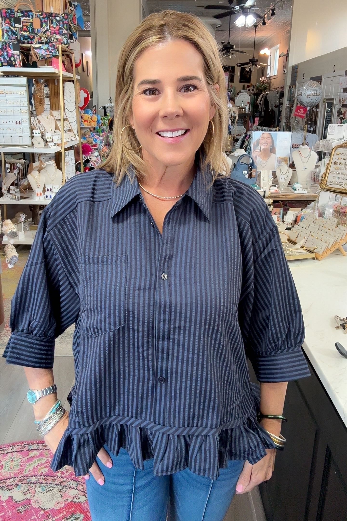 A woman with blonde hair smiling in a boutique shop, wearing a dark navy blue and black vertically striped button-down shirt with ruffle details at the sleeves and hem, paired with blue jeans.