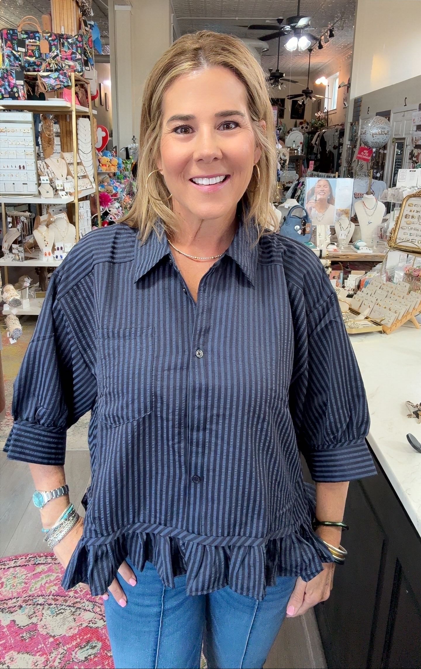 A woman with blonde hair smiling in a boutique shop, wearing a dark navy blue and black vertically striped button-down shirt with ruffle details at the sleeves and hem, paired with blue jeans.