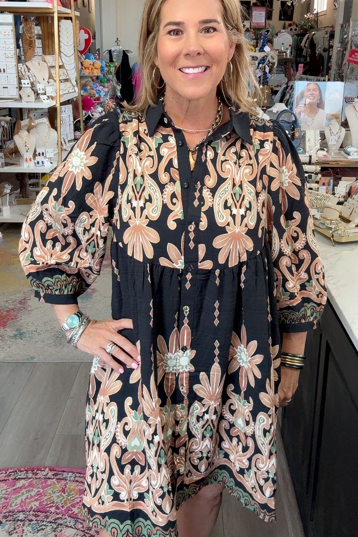 A woman modeling a black button-up dress with beige and pink floral patterns, standing inside a retail boutique.