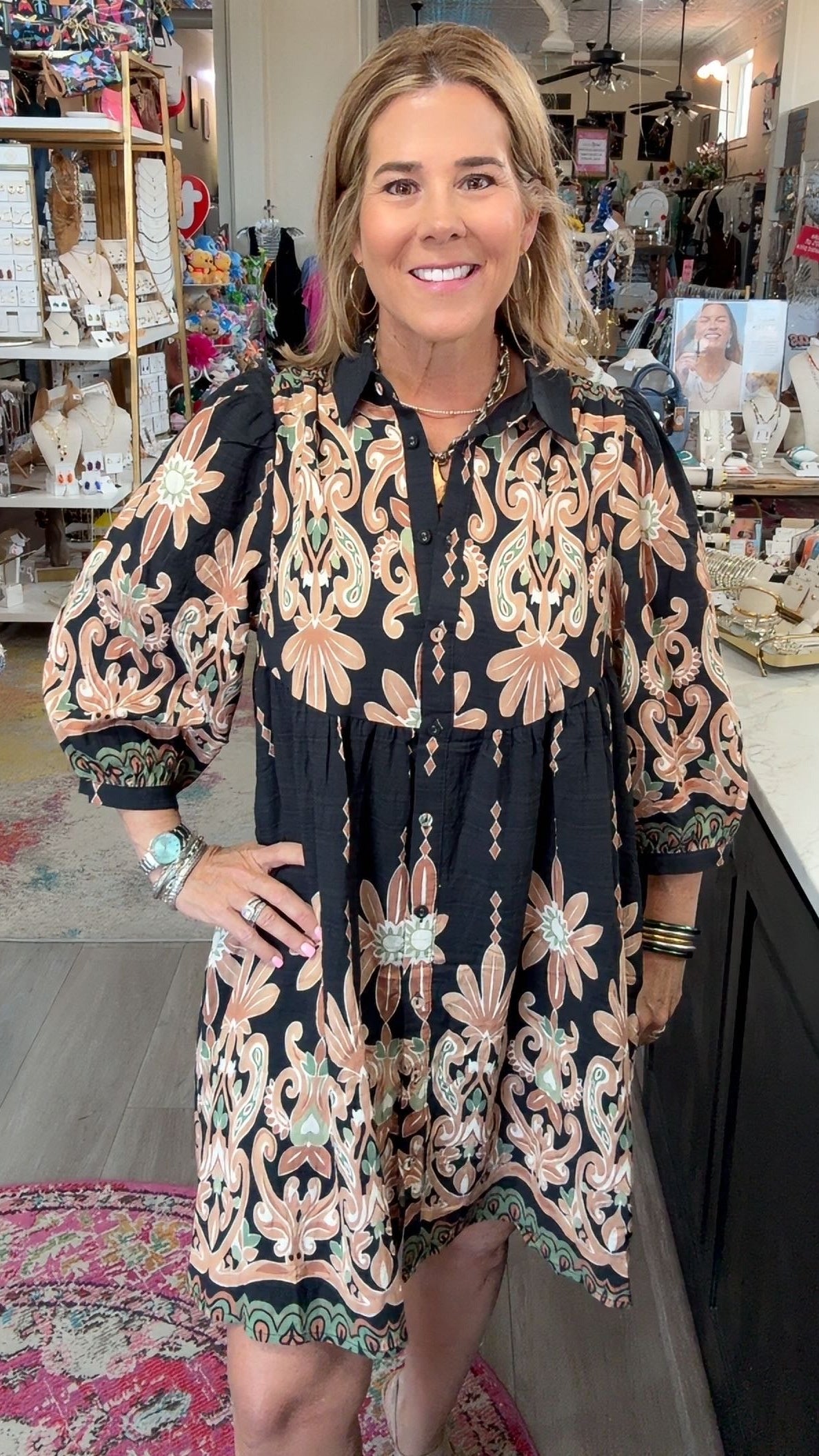 A woman modeling a black button-up dress with beige and pink floral patterns, standing inside a retail boutique.