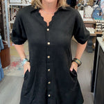 A woman wearing a black short-sleeve button-down shirt dress with scalloped edges and front pockets, standing inside a retail boutique.