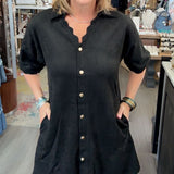 A woman wearing a black short-sleeve button-down shirt dress with scalloped edges and front pockets, standing inside a retail boutique.