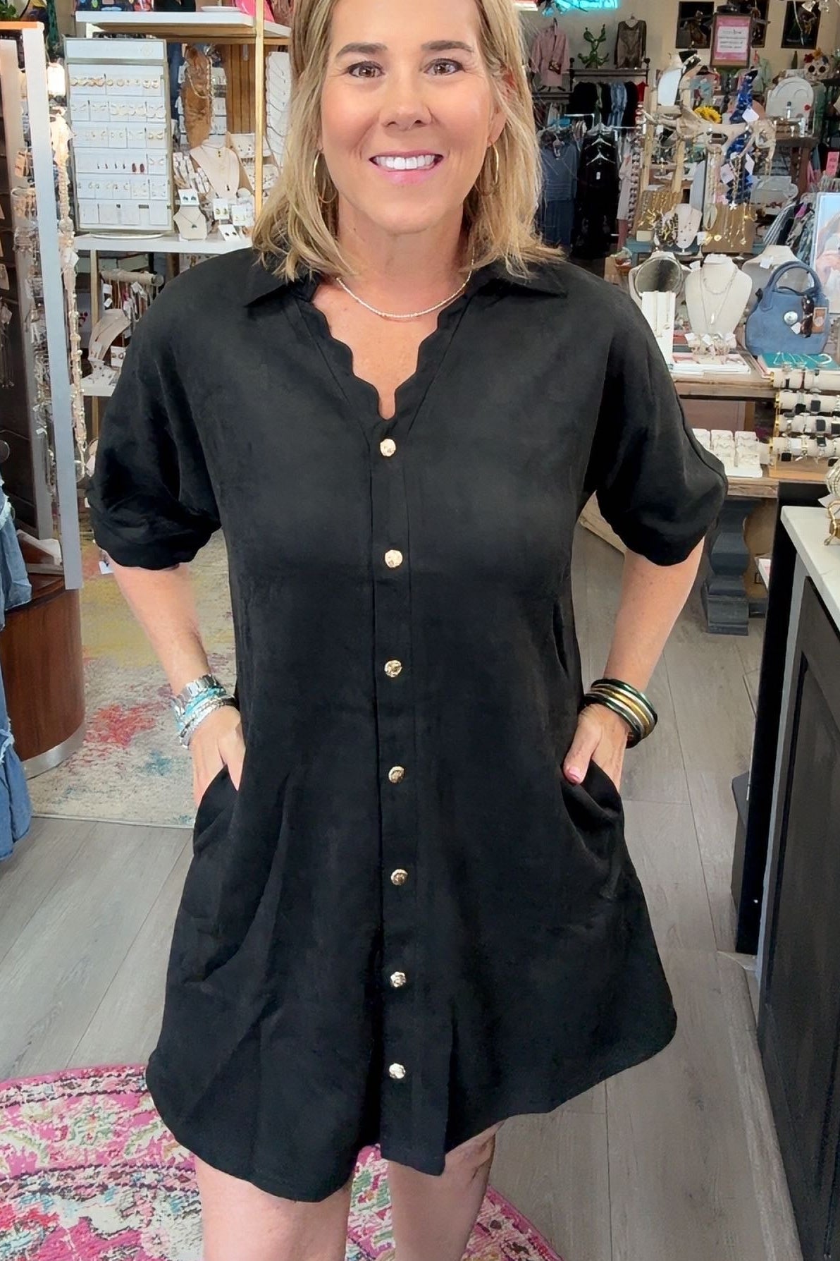 A woman wearing a black short-sleeve button-down shirt dress with scalloped edges and front pockets, standing inside a retail boutique.