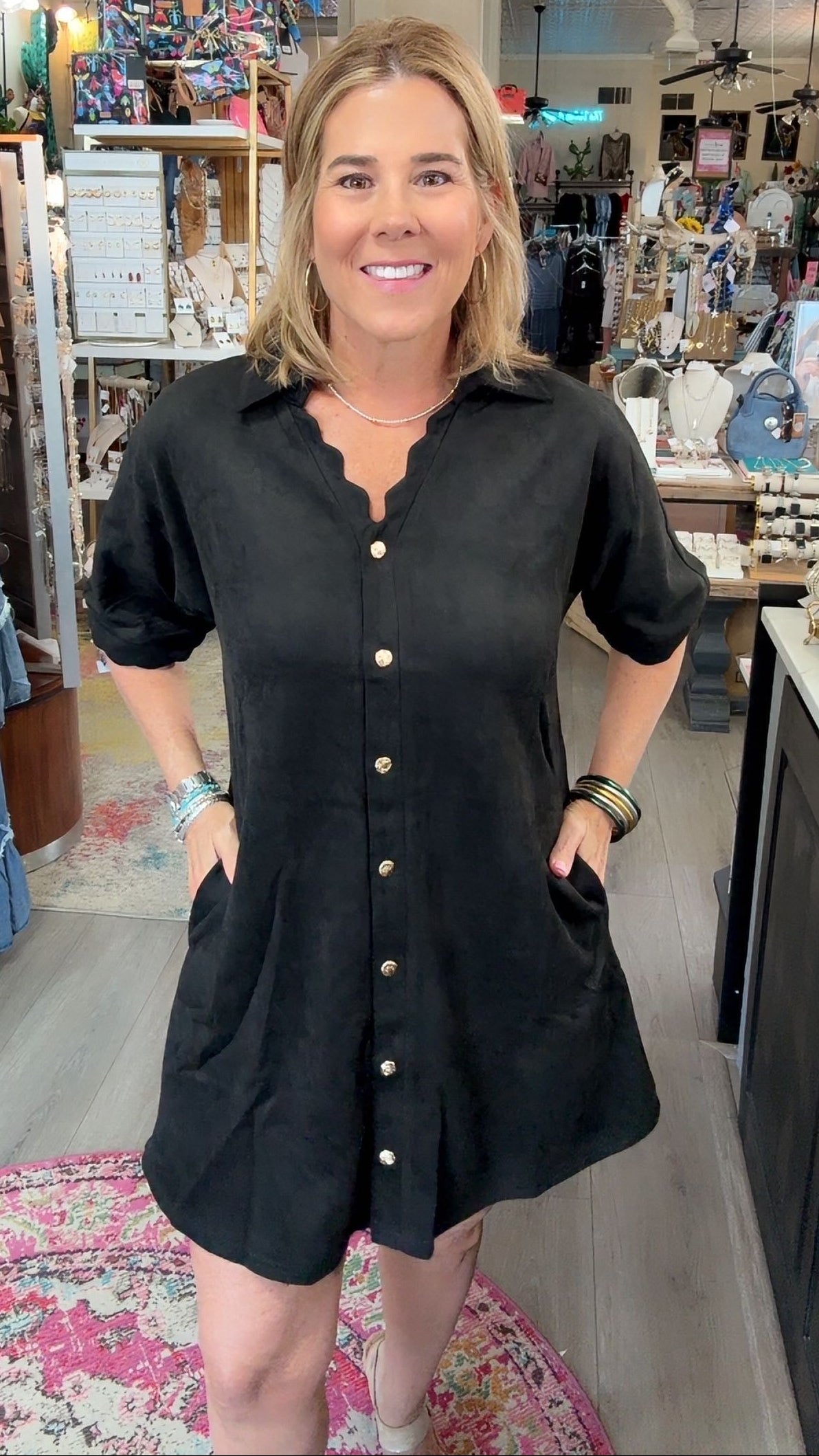 A woman wearing a black short-sleeve button-down shirt dress with scalloped edges and front pockets, standing inside a retail boutique.