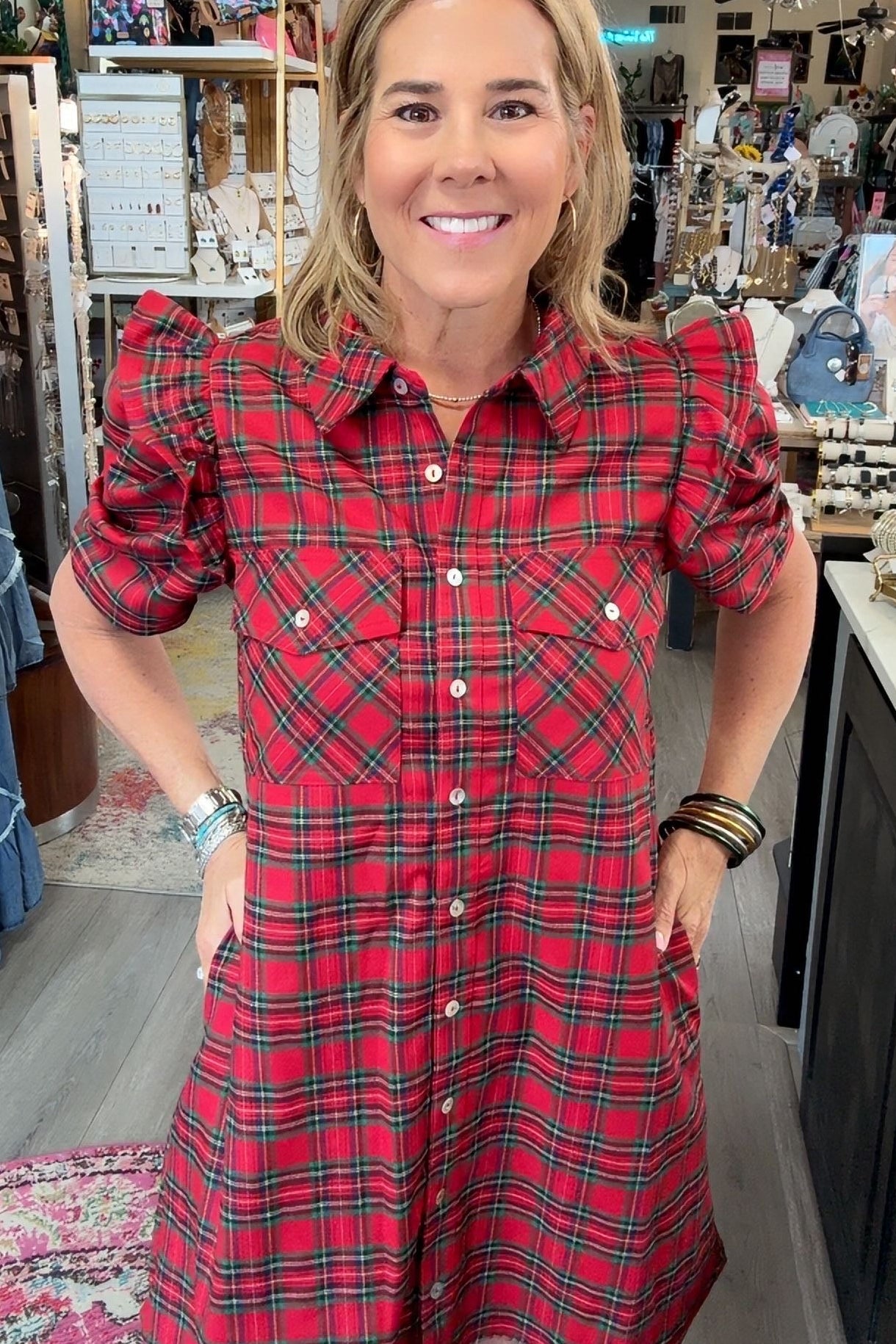 A woman modeling a red and green plaid puff-sleeve shirt dress inside a boutique store.
