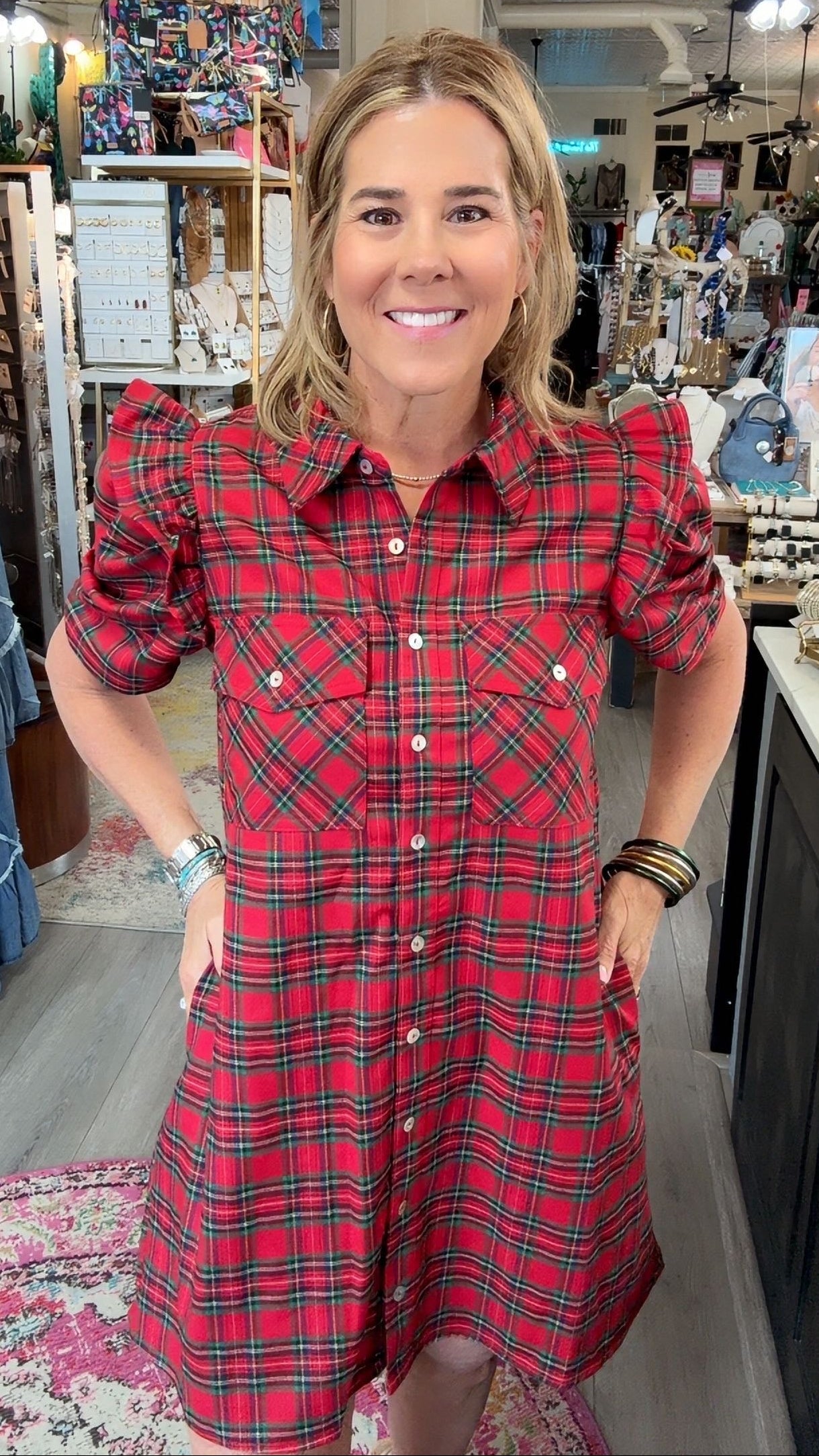 A woman modeling a red and green plaid puff-sleeve shirt dress inside a boutique store.