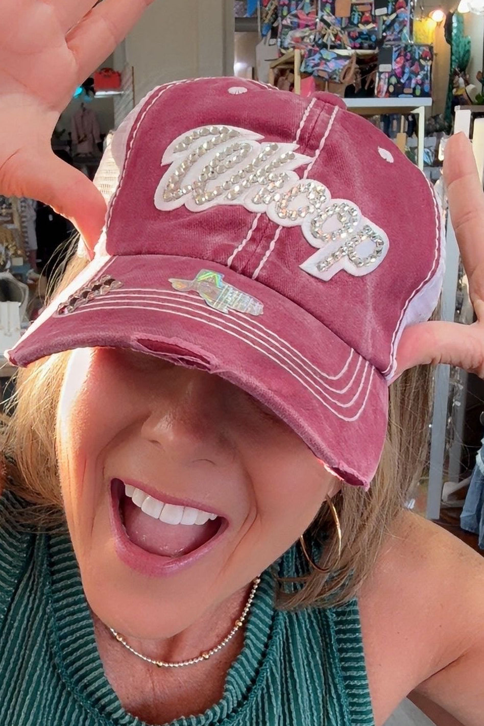 A woman in a green ribbed top posing with a distressed dusty rose baseball cap that has white embroidered text reading 'Crazy Exes Love'. She is smiling and making a gesture with her hands inside what appears to be a retail store.