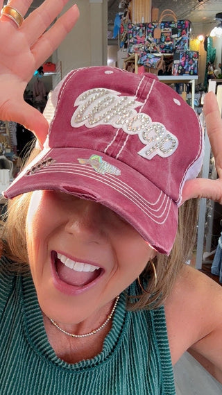 A woman in a green ribbed top posing with a distressed dusty rose baseball cap that has white embroidered text reading 'Crazy Exes Love'. She is smiling and making a gesture with her hands inside what appears to be a retail store.