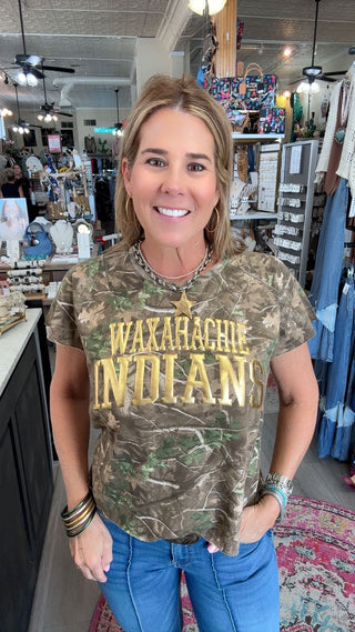A woman wearing a camouflage graphic t-shirt with 'WAXAHACHIANS INDIANS' printed in gold lettering, standing inside a retail store.