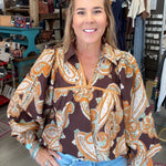 A woman wearing a brown paisley blouse with orange and white patterns, paired with light blue jeans, standing inside a retail store.