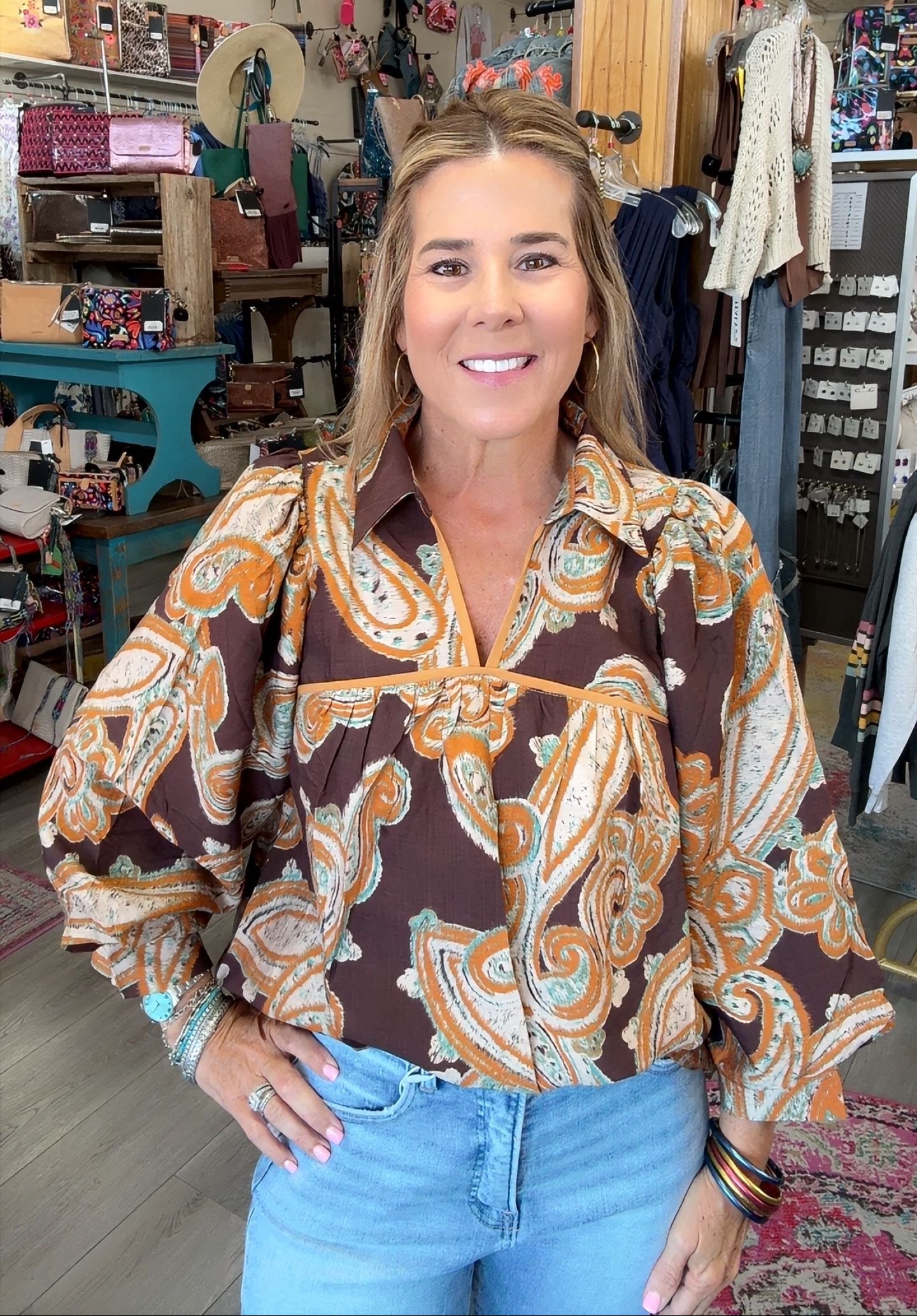 A woman wearing a brown paisley blouse with orange and white patterns, paired with light blue jeans, standing inside a retail store.