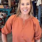 A smiling woman with blonde hair stands in a retail store wearing a textured terracotta orange blouse with puff sleeves and light blue jeans.