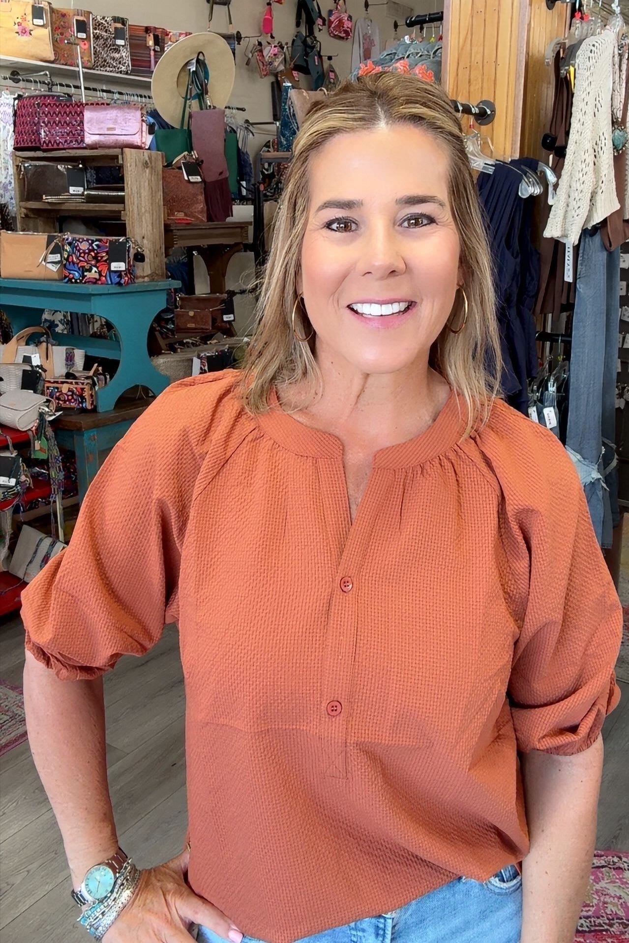 A smiling woman with blonde hair stands in a retail store wearing a textured terracotta orange blouse with puff sleeves and light blue jeans.