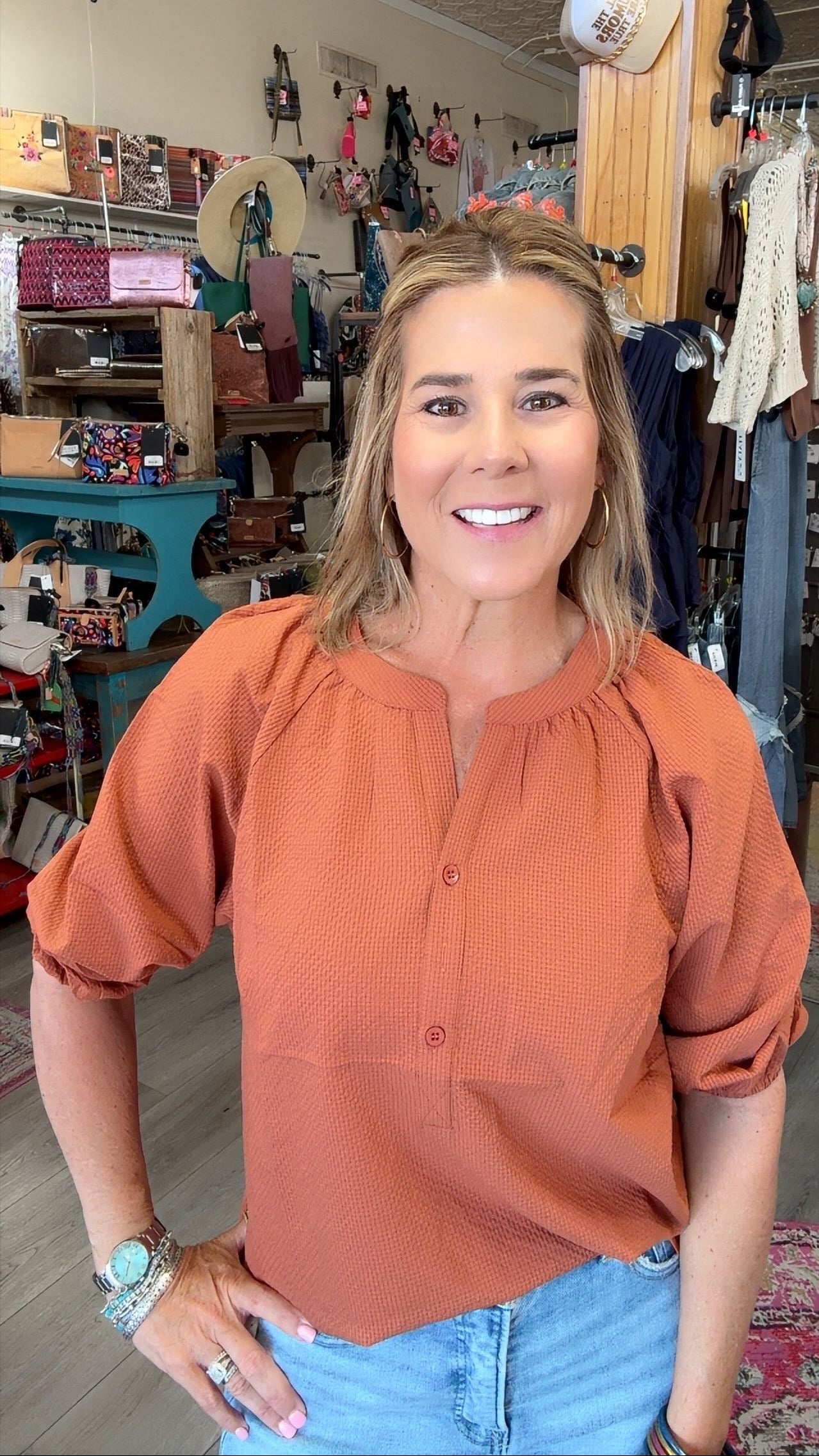 A smiling woman with blonde hair stands in a retail store wearing a textured terracotta orange blouse with puff sleeves and light blue jeans.