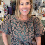 A woman smiling in a boutique shop, wearing a short-sleeve puff-sleeve top with a brown and dusty pink leopard print, paired with light blue jeans.