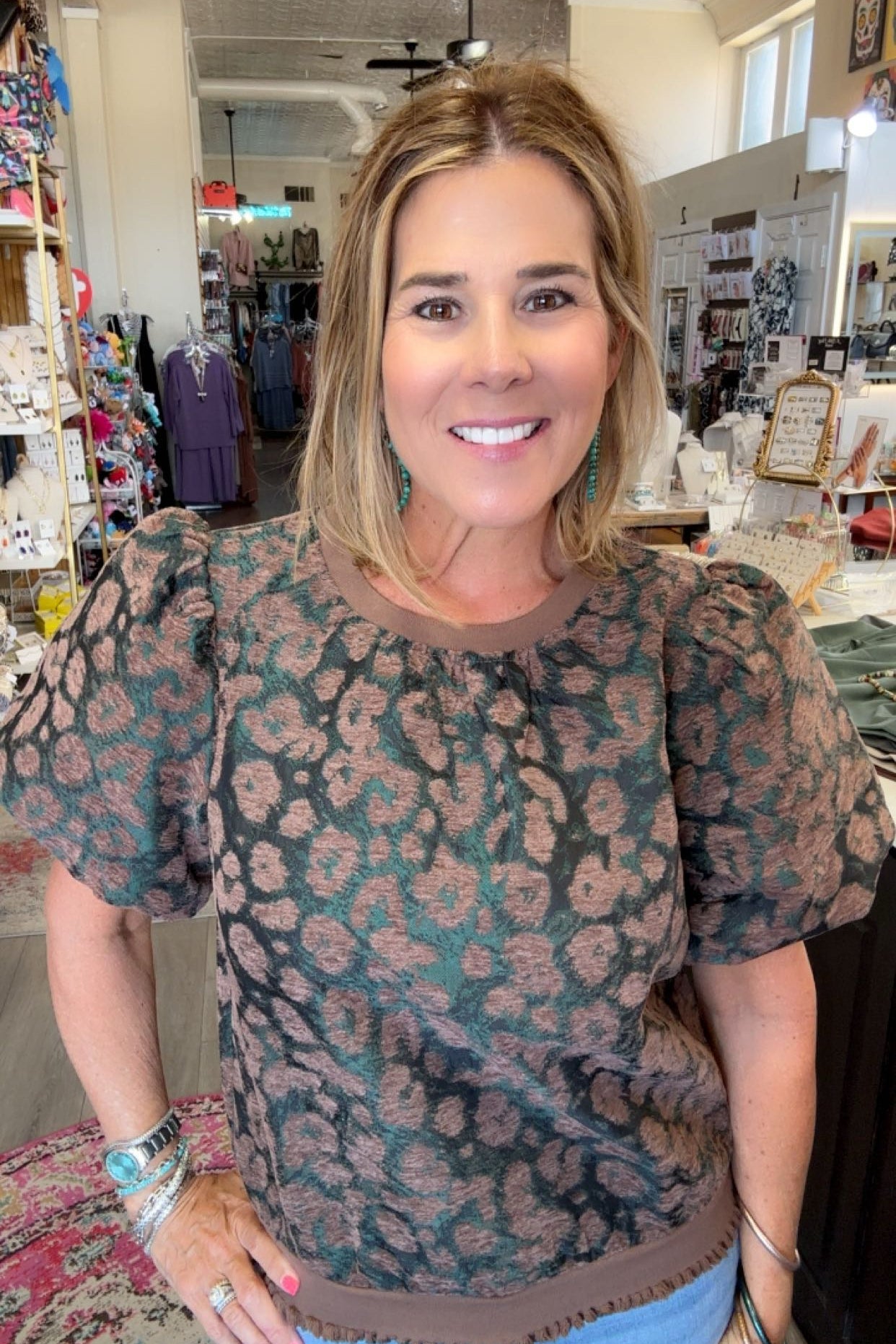 A woman smiling in a boutique shop, wearing a short-sleeve puff-sleeve top with a brown and dusty pink leopard print, paired with light blue jeans.