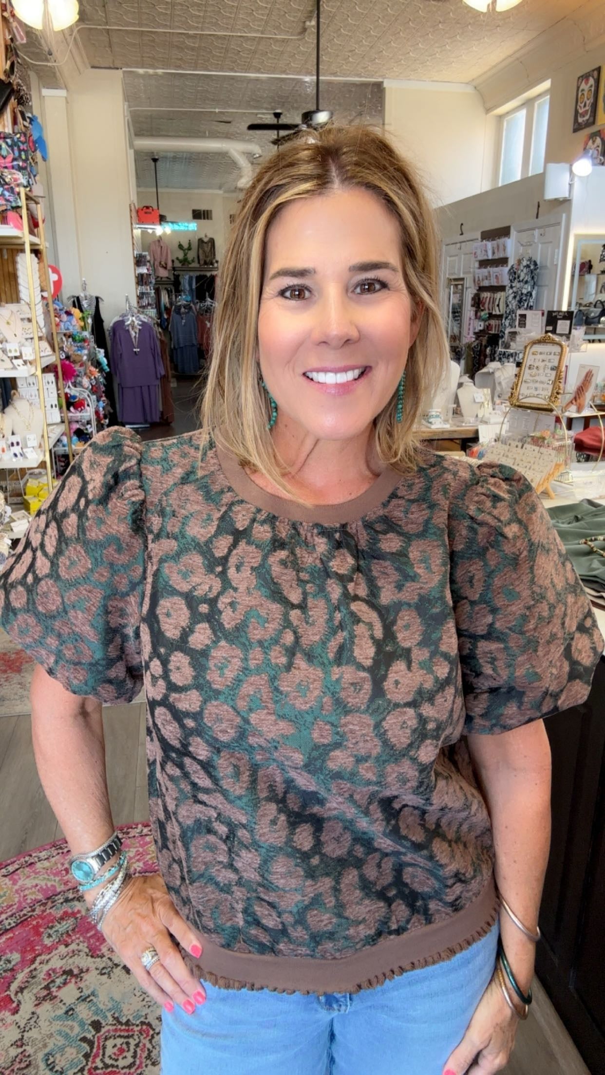 A woman smiling in a boutique shop, wearing a short-sleeve puff-sleeve top with a brown and dusty pink leopard print, paired with light blue jeans.